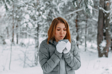 A beautiful girl in a gray jacket and knitted white mittens is drinking a drink in a snowy forest. Comfort and warmth in the winter season. selective focus