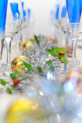 A close-up of a decorated Christmas table, with blue crystal glasses and a gray decorated background with gold garlands and overall silver-colored Christmas outfit.