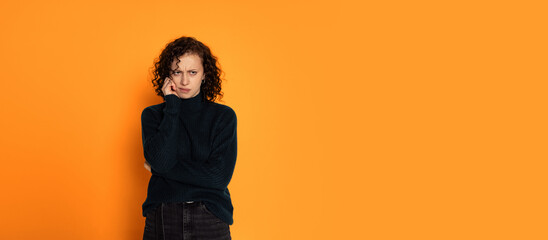 Portrait of young beautiful sad girl in warm knitted sweater is melancholy isolated on orange color background