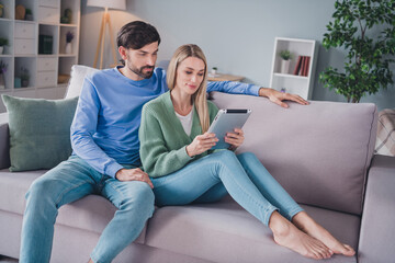 Portrait of two attractive focused people sitting on divan spending weekend using device gadget watching tv video at home indoors