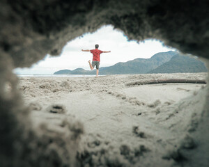 homem correndo de braços abertos na areia da praia, visto a partir de buraco na areia 