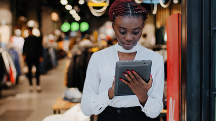 African american serious successful salesperson woman consultant in clothing store shopkeeper boutique employee looks digital tablet makes inventory accounting works online reads discount information - obrazy, fototapety, plakaty
