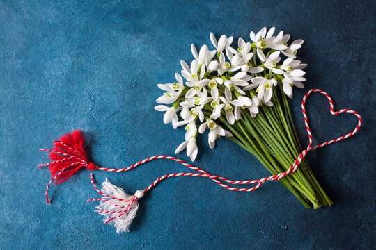 A bouquet of snowdrops flowers and a red and white rope with tassels, a symbol of the arrival of spring on a blue background. Postcard for the feast of March 1 Martisor and Baba Marta, space for text.