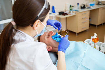 Obraz premium Dentist during the treatment of a male patient. Female dentist in face mask with male patient sitting in dentist chair sitting in dental clinic. Selective focus