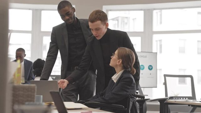 Low angle of young Caucasian and Black men wearing formal attire standing by desk in office, consulting young woman in wheelchair using laptop computer in morning