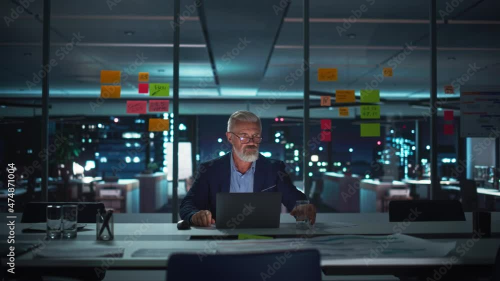 Poster successful senior businessman working on laptop computer in big city office late in the evening. inv