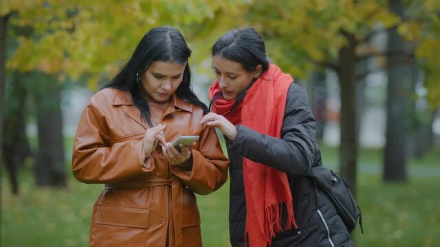 Two beautiful hispanic young women stand in autumn park girls using smartphone look at gadget choosing clothes in internet store shopping online females use cool new phone app play on mobile telephone
