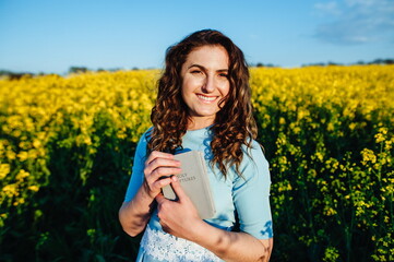 Portrait of a happy female student holding books and looking at camera outdoors. Young, beautiful girl holding a Bible in her hands, against the background of yellow flowers in the field.