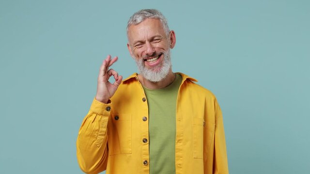 Smiling elderly gray-haired bearded man 50s wears yellow shirt showing okay ok zero fingers gesture isolated on plain pastel light blue background studio portrait. People emotions lifestyle concept