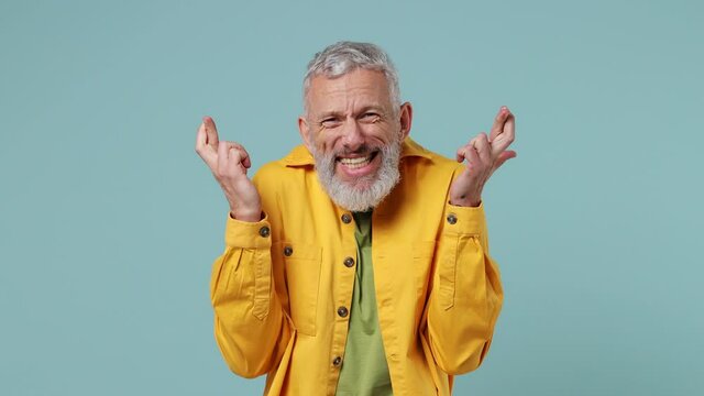 Elderly gray-haired bearded man 50s wears yellow shirt hold hands folded in prayer begging about something make wish keep fingers crossed isolated on plain pastel light blue background studio portrait