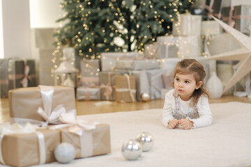 Adorable little girl lying on her front nearby fir tree and smile. Christmas mood at home with lot of presents