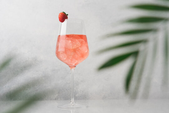 Glass of pink cold cocktail with ice surrounded by palm leaves. Summer refreshing drink on white background, side view, close-up view