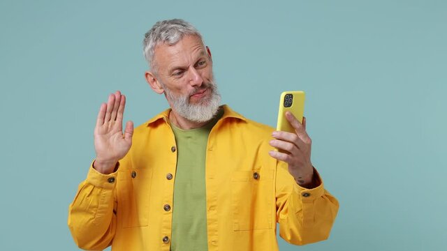 Happy fun elderly gray-haired bearded man 50s wears yellow shirt get video call using mobile cell phone doing selfie talk greet with hand isolated on plain pastel light blue background studio portrait