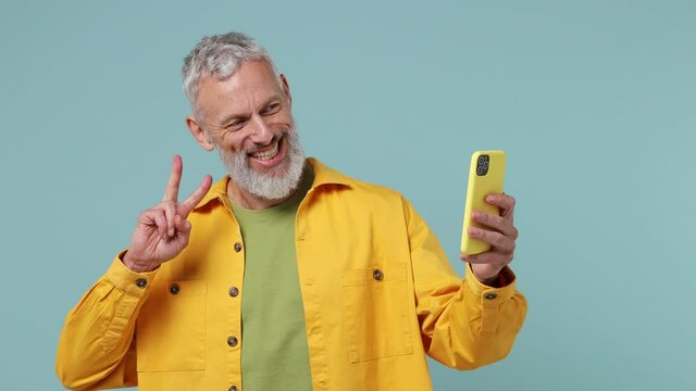 Happy charismatic elderly gray-haired bearded man 50s wears yellow shirt doing selfie shot on mobile phone post photo on social network isolated on plain pastel light blue background studio portrait