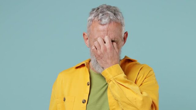 Sick ill tired jaded elderly gray-haired bearded man 50s wears yellow shirt put off glasses rub nose suffer from migraine feel bad seedy isolated on plain pastel light blue background studio portrait