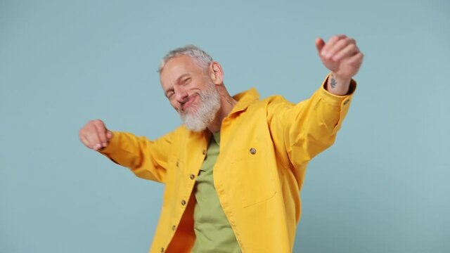Slow motion fancy vivid elderly gray-haired bearded man 50s in yellow shirt dance fool around have fun gesticulate with hands enjoy relax isolated on plain pastel light blue background studio portrait