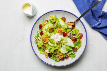 Waldorf salad, made of fresh green apples, celery, walnuts, grapes, dressed in mayonnaise and yogurt sauce, and served with lettuce. Top view, white table surface.