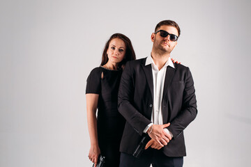 young couple, in black suits with pistols in their hands on a white background in the studio, they depict security guards, bodyguards or agents.