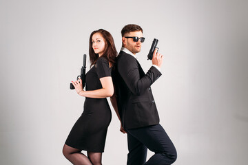 young couple, in black suits with pistols in their hands on a white background in the studio, they depict security guards, bodyguards or agents.