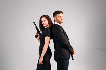 young couple, in black suits with pistols in their hands on a white background in the studio, they...