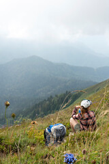 Young woman traveler in plaid shirt in defocus sitting on meadow in mountain valley next to backpack drinking water from bottle on hike mountain landscape with blue flowers in foreground