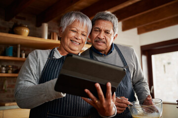 Multi-cultural elderly couple smiling, using tablet to research recipe in modern kitchen. Happy couple cooking healthy meal.