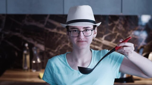 portrait of a teen ia a hat preparing dinner at home. happy teen tasting soup on spoon in kitchen and smiling. pretty girl prepares soup on the stove in the kitchen.