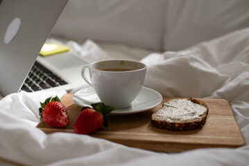 Morning coffee in bed. Cup of fragrant, aromatic coffee, sandwich and two ripe strawberries on wooden kitchen board lies near laptop on white bed. Beakfast at home-office in time quarantine.