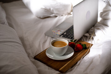 Morning coffee in bed. Cup of fragrant, aromatic coffee and two ripe strawberries on wooden board near laptop on white bed. Beakfast at home-office in time quarantine.