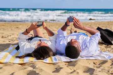 Young multicultural couple lying on the beach using smartphones