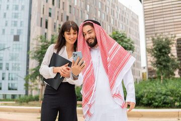 Smiling arab businessman showing video on smartphone to caucasian businesswoman at meeting outdoors
