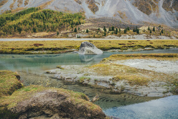 Golden autumn landscape with big stone in mountain creek with view to forest hillside. Beautiful big boulder in turquoise clear water stream in autumn. Green ripple sandy bottom in transparent water.