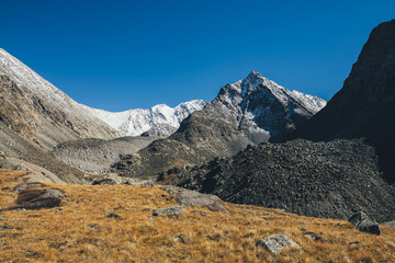 Autumn landscape with highland valley among snow-covered mountain range and pointy peak under blue sky. Atmospheric mountain scenery with sunlit orange valley in autumn colors among rocky mountains.