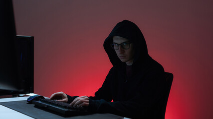 Isolated Handsome Caucasian Hacker Man Sitting In Front of the Computer, Hands on the Keyboard, Background is Red Colored, Hacking Into the Servers, Conducting an Attack on Social Media Platforms.
