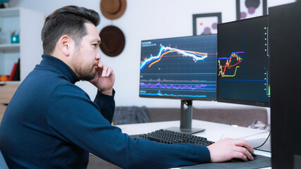 Handsome Asian Stock Broker Working Remotely From Home. Looking Focused and Serious As He Works Behind Two Computer Screens.