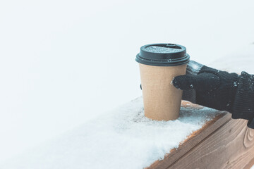 One disposable coffee cup on the snow on a wooden board. A hand in a knitted black glove holds a package