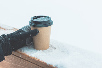 One disposable coffee cup on the snow on a wooden board. A hand in a knitted black glove holds a...