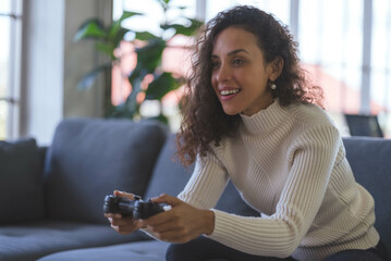 Young African American woman is having fun playing video games with a single joystick on sofa at home. Social distancing during the COVID-19 epidemic. © amornchaijj