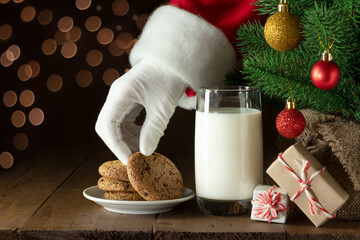 Santa Claus hand takes chocolate chip cookie. Snacks and glass of milk for Santa under Christmas...