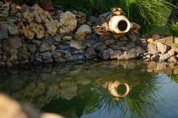 Naklejka premium The decorative pitcher is reflected in the artificial pond. Beautiful summer photo of decorative garden decoration close-up.