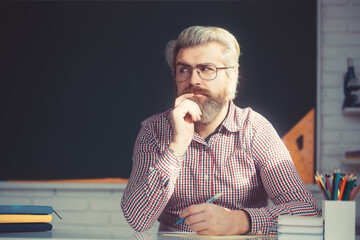 Attractive male teacher sitting thinking at his desk with his hand resting on a pile of textbooks.