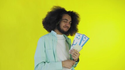 A happy African-American man looks at the camera, holds tourist vouchers in his hand, stands isolated on a yellow background