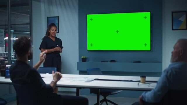 Company Operations Manager Holds Meeting Presentation for a Team of Executives. Multiethnic Female Uses Wide TV Set with Horizontal Green Screen Mock Up Display. People Work in Business Office.
