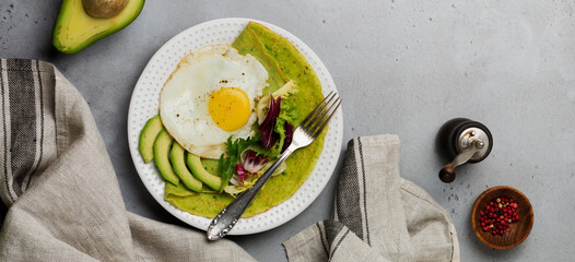 Spinach green crepes (pancakes) with fried egg, avocado and leaves of mix of salad on ceramic plate on gray concrete background. Concept of healthy breakfast. Selective focus. Top view. Copy space.