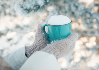 Winter image of woman's hands in the wool mittens holding vintage blue mug full of clear white snow. Winter freshness concept. Awesome snowy forest in sunny winter morning. Winter weekend out of town.