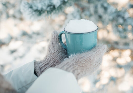 Winter image of woman's hands in the wool mittens holding vintage blue mug full of clear white snow. Winter freshness concept. Awesome snowy forest in sunny winter morning. Winter weekend out of town.