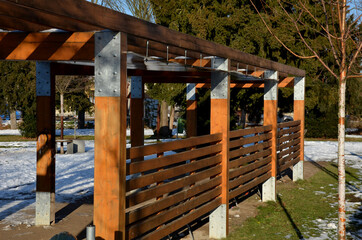 behind a hedge in the park stands a new pergola made of brown wood by a landscape architect. is...