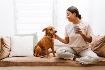 Young hispanic woman petting her dog while drinking tea
