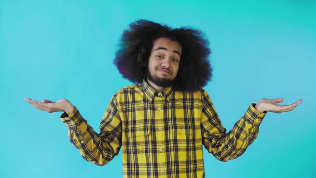 A young man with an African hairstyle on a blue background shrugs his shoulders. Emotions on a colored background.