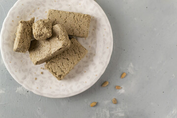 Several pieces of sunflower halva on a white saucer with seeds on a gray background. Concept of traditional oriental sweets. Horizontal orientation. Top view. Copy space.